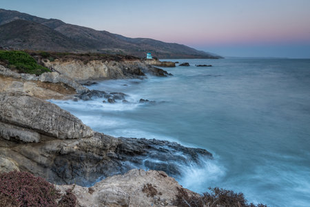 Waves crash against rocky cliffs at Leo Carrillo State Beach during twilight, overlooking the tranquil ocean and distant mountains in Malibu.の写真素材