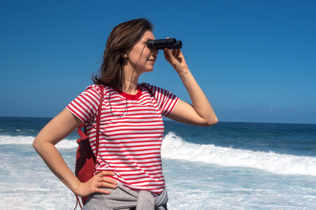 Girl tourist looking through binoculars on background of oceanの写真素材