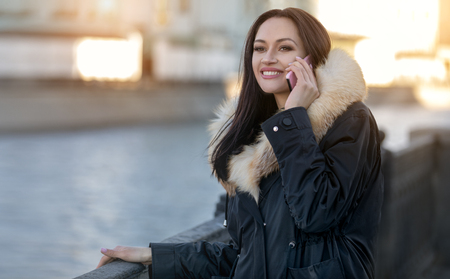 A girl in a warm jacket with a fur collar on the street talking on the phone. Cold autumn weather and street fashionの写真素材