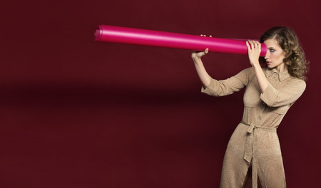 girl model looks away through the roller twisted of pink plastic. fashion shooting in Studio on dark red background. The model is holding a telescope.の写真素材