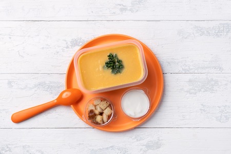 soup puree in a plastic box on a plastic orange plate. plate stands on a white wooden table. fast food and crackers and sour cream in a box. the view from the topの写真素材