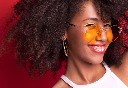 smiling model with dark curly hair in orange glasses on a red background of the Studio. smiles and looks playfully. There is a place for copy pasteの写真素材