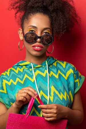 dark-skinned girl with lush curly hair posing with shopping bags on red Studio background. the girl glasses with starsの写真素材