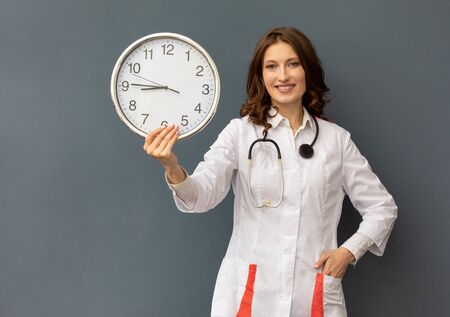 a woman doctor with a stethoscope around her neck and a large wall clock in her hands. the concept of time that curesの写真素材