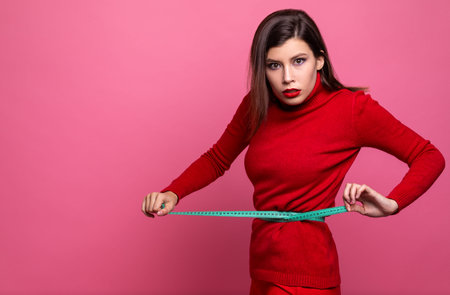 a girl in a red sweater and red pants measures her waist with a centimeter tape measure. girl...