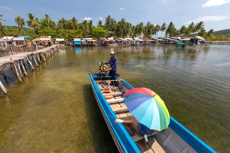 Vietnam Phu Quoc island 2 April 2019. Vietnamese boat driver with a motor carries a tourist with a bright umbrella. in the background behind them is a fishing villageのeditorial素材