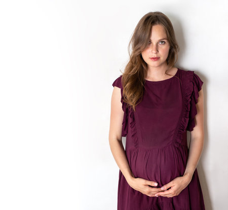 a pregnant girl stands against a white wall. hands holds its big stomach
