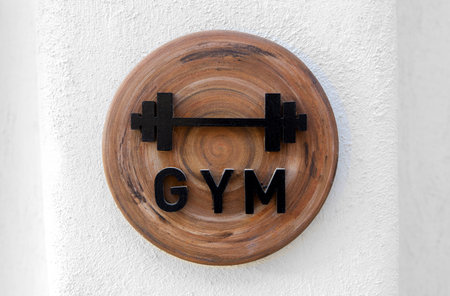 A wooden circular sign with a black barbell icon and the word "GYM" in bold letters. The sign is mounted on a white wall, symbolizing a fitness center or gym environmentの写真素材