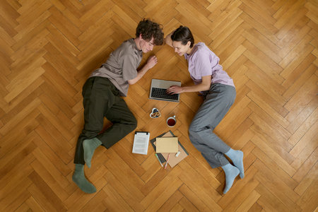 Top view of young caucasian couple in love smiling laughing looking at each other lying on the wooden floor and watching films using laptop while eating snacks in the living room.の写真素材