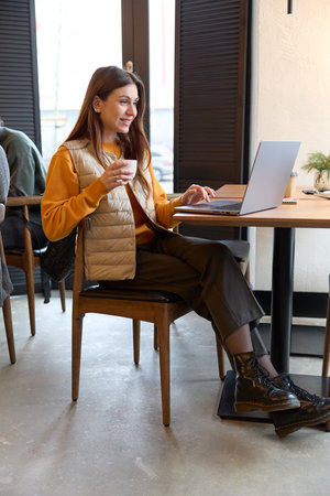 Vertical side shot in full height of a young student girl working on a computer doing assignment and holding a cup of coffee in her hand while sitting on a chair at a table.の写真素材