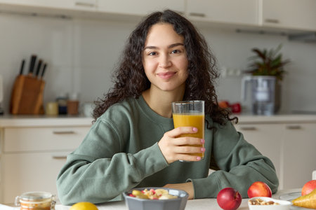 Portrait of young curly beautiful smiling girl looking at camera wearing green sweater having a healthy breakfast and holding a glass of freshly squeezed juice eating granola.の写真素材