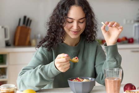 Close up of beautiful young curly haired smiling woman having a healthy breakfast holding a spoon eating granola with fruits and drinking smoothie while sitting at a kitchen table.の写真素材