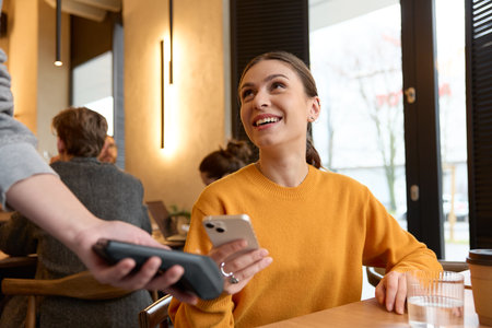 Portrait of a female client smiling and using smartphone to pay for coffee to crop waiter while sitting at table in the morning in a light cafe indoors. Lifestyle conceptの写真素材