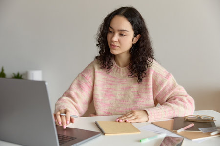 Close up of young beautiful curly haired focused student girl preparing for exam holding a pen writing in her notepad and working on the computer while sitting at a table in a niceの写真素材