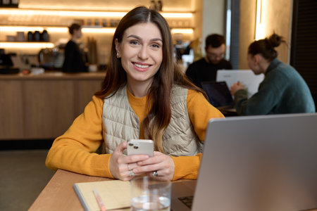 Horizontal shot of beautiful caucasian female smiling dressed in casual yellow sweater and beige vest using a smartphone working on laptop looking at camera posing while sitting.の写真素材