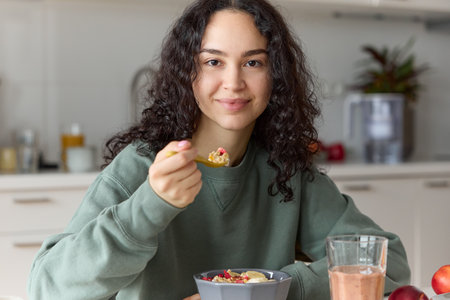 Close up of pretty young curly smiling woman looking at camera having a healthy breakfast holding a spoon eating cereal muesli with berries and drinking smoothieの写真素材