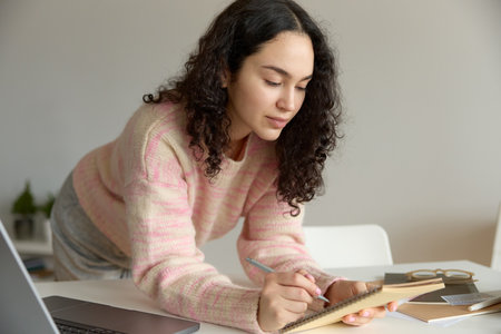 Close up of young beautiful curly haired woman wearing pink sweater writing making notes in a notebook while working on a laptop at her kitchen table at home. Lifestyle concept.の写真素材