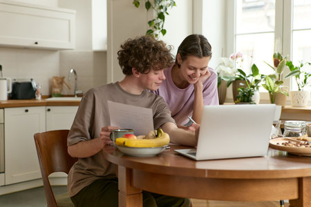 Portrait of young beautiful happy smiling european couple taking care of finance reviewing documents calculating bills using laptop sitting at dining table with papers.の写真素材