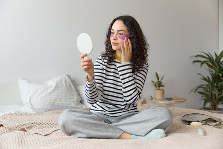 Portrait of a young beautiful curly haired woman wearing gel purple patches under eyes and looking in a little mirror while sitting on a bed with her legs crossed.の写真素材