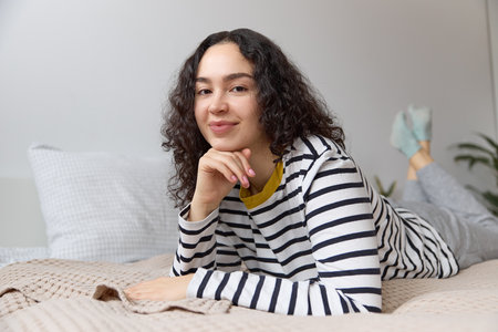 Close up of a young beautiful woman with long curly brown hair wearing striped pajama smiling looking at camera lying relaxing on a bed in a modern light apartment indoors.の写真素材