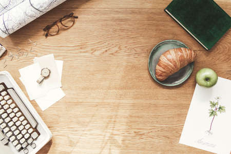 Stylish composition of flat lay on wooden desk with croissant, map, floral poster, notebook and...