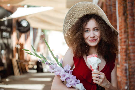 Summer sunny lifestyle portrait of young beautiful woman walking on the street market with flowers bouquet, wearing cute red trendy dress, eating icecream, smiling enjoy her holiday.の写真素材