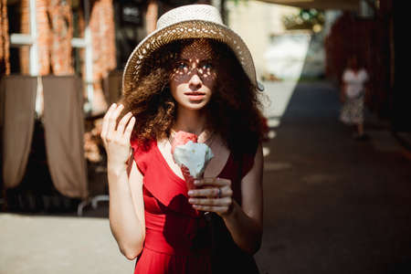 Sunny lifestyle portrait of young beautiful woman walking on the street market, wearing cute red trendy dress, eating icecream, smiling enjoy her holiday.の写真素材