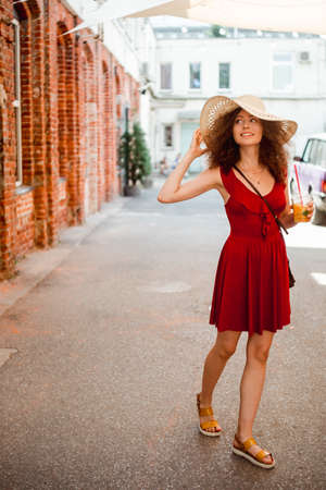 Summer sunny lifestyle portrait of young beautiful woman walking on the street market, wearing cute red trendy dress, drinking tasty smoothie, smiling enjoy her weekends, holiday.の写真素材
