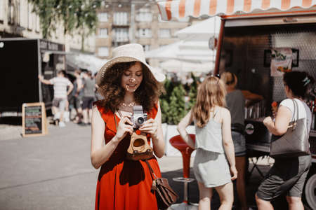 Summer sunny lifestyle portrait of young beautiful woman walking on the street market with photo camera, wearing cute red trendy dress, smiling enjoy her weekends, holiday.の写真素材