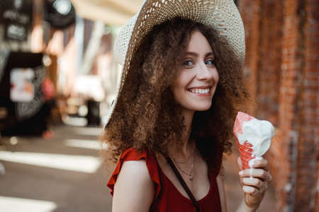 Sunny lifestyle portrait of young beautiful woman walking on the street market, wearing cute red trendy dress, eating icecream, smiling enjoy her holiday.の写真素材