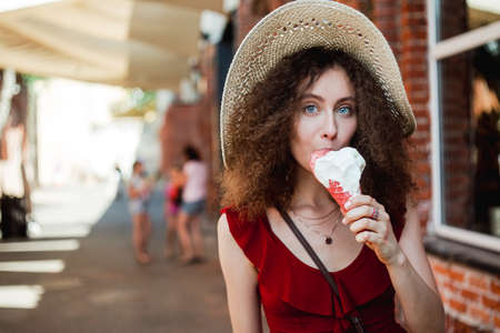 Sunny lifestyle portrait of young beautiful woman walking on the street market, wearing cute red trendy dress, eating icecream, smiling enjoy her holiday.の写真素材