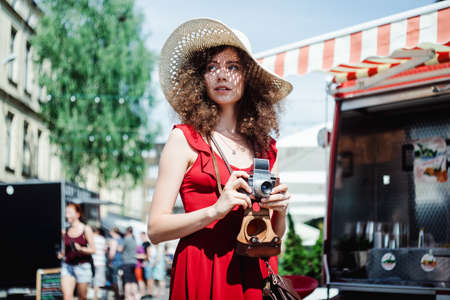 Summer sunny lifestyle portrait of young beautiful woman walking on the street market with photo camera, wearing cute red trendy dress, smiling enjoy her weekends, holiday.の写真素材