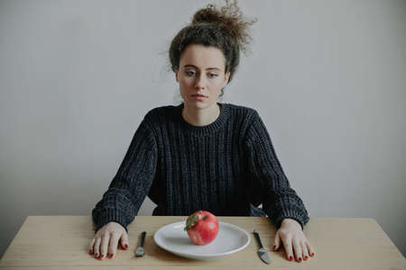 Young woman with anorexia and depression sitting opposite the kitchen table with plate. Sadness,...