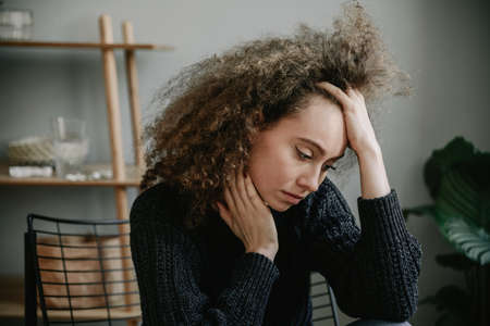 Dark hair teenager with depression sitting alone on the armchair in gray room. Sadness, nostlagic, depression. Mental problems of young people.の写真素材