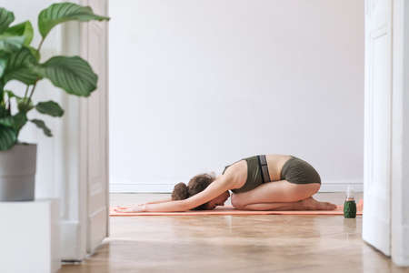 Young darkhair woman practicing yoga in the morning at her home. Big open space, white walls, brown...