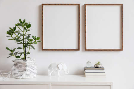 Stylish white home decor of interior with two brown wooden mock up photo frames with books, beautiful plant in stylish pot, elephant figure and home accessories. Minimalistic scandinavian room.の写真素材