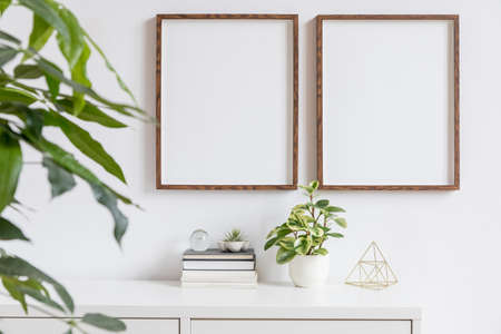 Stylish home interior with two brown wooden mock up photo frames on the white shelf with books, beautiful plants, gold pyramid and home accessories. Minimalistic concept of white room decor.の写真素材