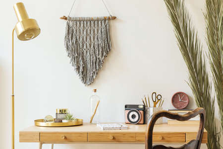 Stylish scanidnavian interior with wooden desk, grey macrame, design accessories, photo camera, gold lamp and wooden ladder on the white wall. Piece of design woman's room. Creative desk of home decorの写真素材
