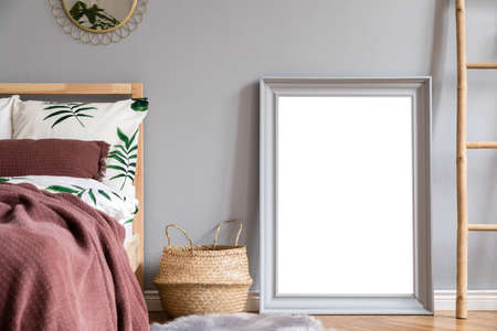 Stylish and luxury interior of bedroom with gray mock up frame, gold mirror, wooden ladder and elegant accessories. Beautiful floral bed sheets, blankets and pillows. Modern home decor.の写真素材