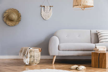 Design bohemian composition of living room interior with gray sofa, wooden cube, beige macrame, hat, rattan lamp and basket with plaid and elegant accessories. Stylish home decor. Template. Gray walls.の写真素材