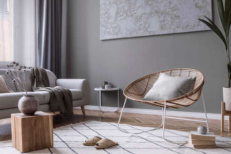 Modern and bohemian composition of interior design with gray sofa, rattan armchair, wooden cube,...