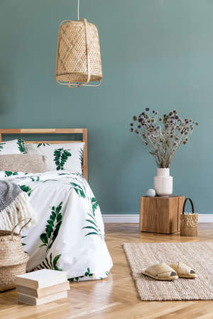 Minimalistic and boho interior design of bedroom with wooden cube, vase with flowers, rattan basket, books and elegant accessories. Beautiful bed sheets, blankets and pillows. Modern home decor.の写真素材