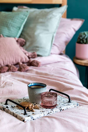 Stylish composition of bedroom interior with lastrico tray with coffee and elegant personal accessories. Beautiful pink and green bed sheets, blanket and pillow. Template Design home staging. Close upの写真素材