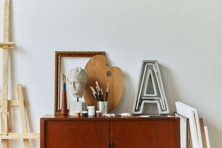 Stylish composition of artist workspace room with design retro teak commode, poster frames, easel, book, decoration and painting accessories. Template.の写真素材