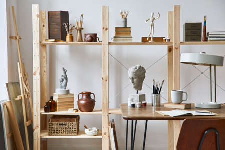 Unique artist workspace interior with stylish desk, wooden easel, bookcase, artworks, painting...