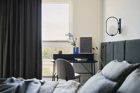 Stylish composition of small modern workspace interior with fposter frame and personal accessories. Bed with linen bedclothes and creative designed lamp in the foreground. Neutral walls with black panels. Panoramic windows with gray curtains. Minimalistic masculine concept. Template.の写真素材