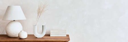 Minimalist composition of living room with wooden commode, table lamp, dried flower in vase, book,...