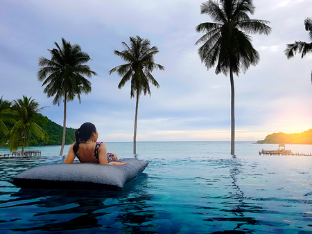 Young woman sit in beanbag relaxing on the swimming pool and looking forward, near the sea in the sunsetの写真素材