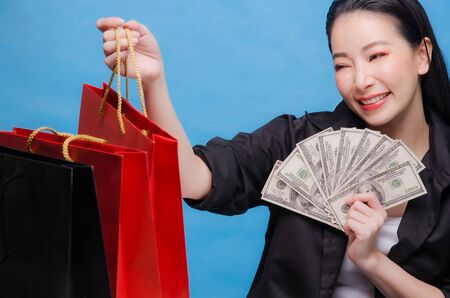 Portrait of a happy Chinese woman in black shirt holding red shopping bag and money isolated on a blue backgroundの写真素材
