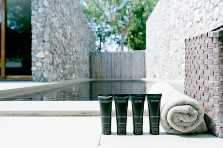 (Blank Label) Mini plastic tube with cosmetic products and towels and basket beside swimming pool against sun light. Hotel amenities such as hair shampoo, hair conditioner, body wash and body lotionの写真素材